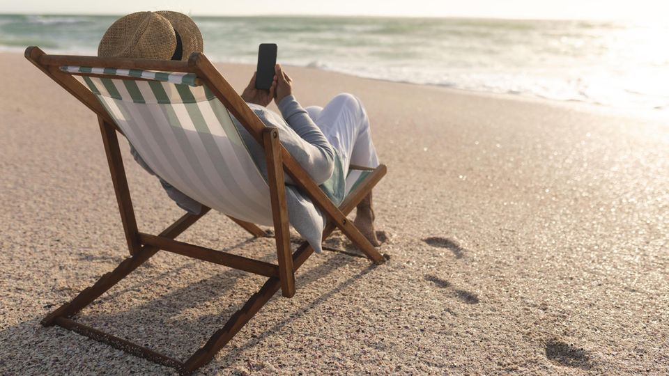 A person is lying in a deck chair on the beach with a smartphone