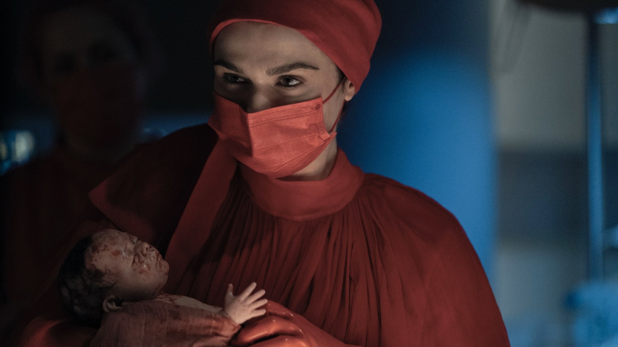 A woman in red scrubs and a red hospital mask cradles a bloody newborn baby.