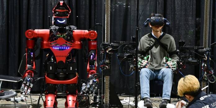 a red humanoid robot on a wheeled platform waits beside a human wearing a vr headset with his hands and arms covered in mechanic.jpg