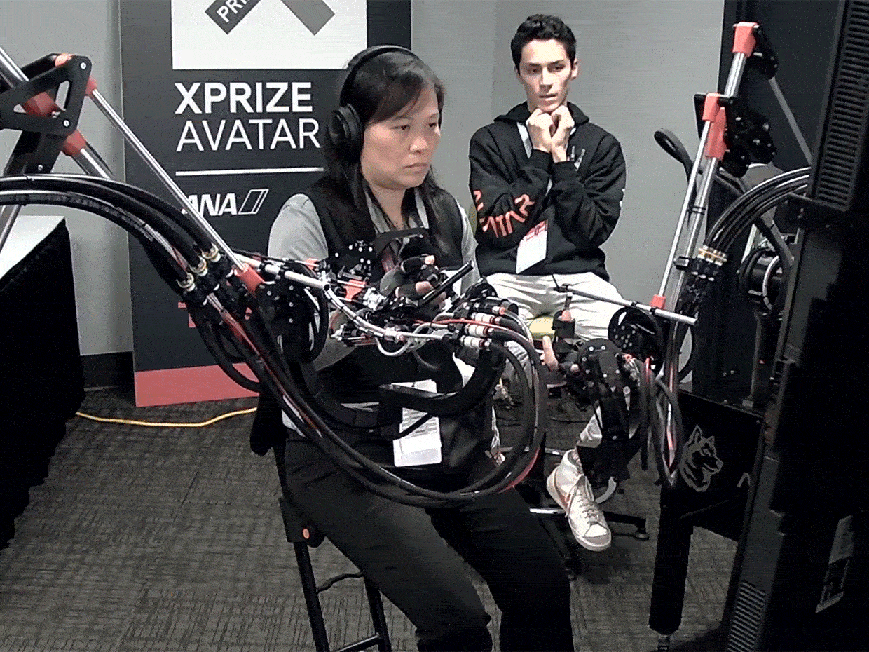A woman wearing robotic gloves controls a remote robot while a man behind her watches anxiously. 
